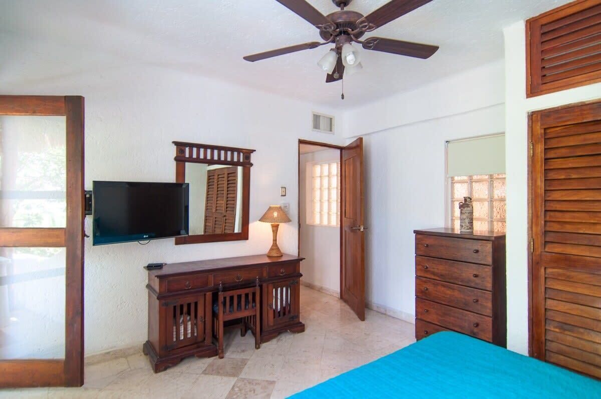A bedroom with a ceiling fan, a TV mounted on the wall, a dresser, a bed with a blue bedspread, and a door leading to another room.