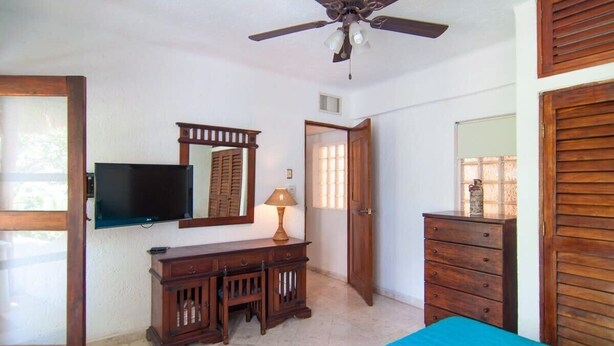 A bedroom with a ceiling fan, a TV mounted on the wall, a dresser, a bed with a blue bedspread, and a door leading to another room. - Private Terrace & Hammock Near Mamitas Beach (Playa del Carmen, Q.R.)
