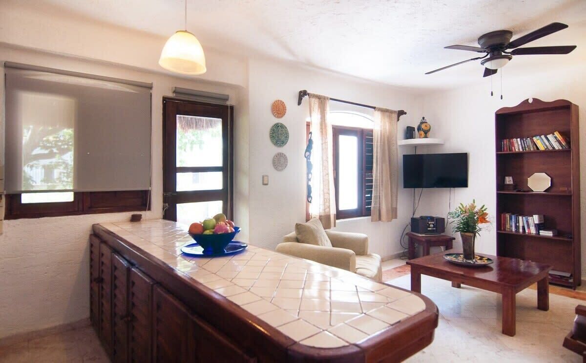 A living room with a sofa, a coffee table, a TV, a bookshelf, and a kitchen area with a counter and cabinets.