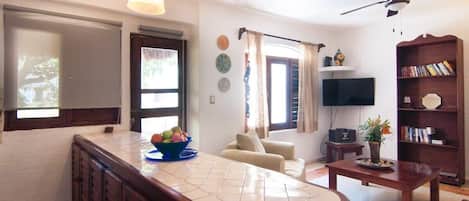 Ein Wohnzimmer mit einer Couch, einem Couchtisch, einem Fernseher, einem Bücherregal und einer Küchenzeile mit Arbeitsplatte und Schränken.