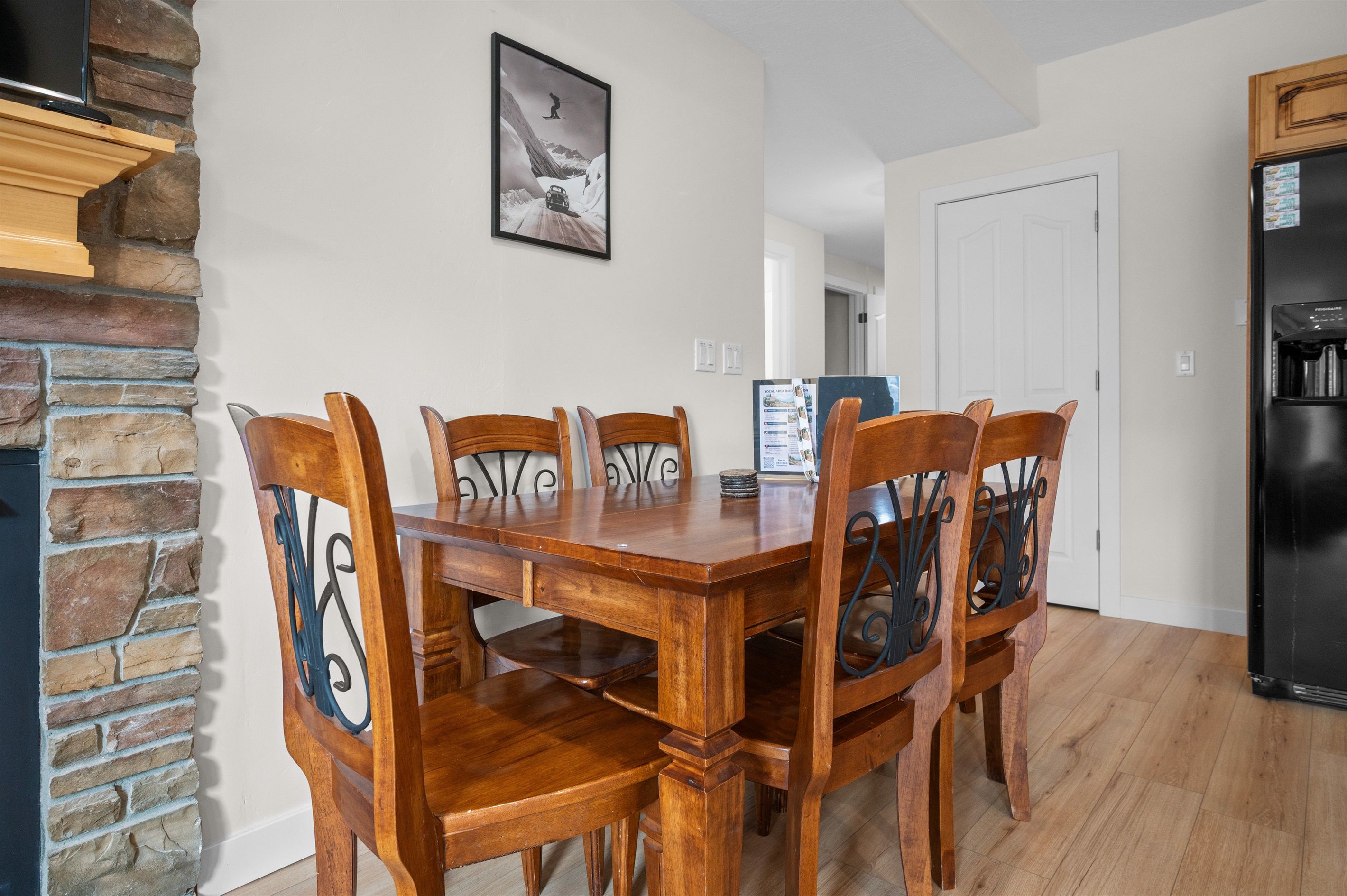 A dining area with a wooden table and chairs, a stone wall, a black refrigerator, and a door in the background.