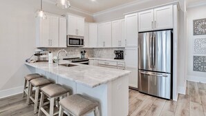 A modern kitchen with white cabinets, a stainless steel refrigerator, a marble island with bar stools, and pendant lighting. - West Side Cottages GG | Beach Cottage, Pet Friendly, Pool, Beach/Lagoon Access! (Gulf Shores)