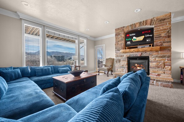 A living room with a stone fireplace, a large sectional sofa, and a flat-screen TV mounted above the fireplace displaying various streaming services. - Lakeside Escape LS36 |Ski Lodge |Hot Tub | Game Rm (Huntsville)