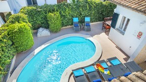 Una piscina con sillones de descanso y una mesa, rodeada de follaje y un techo de baldosas.