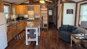 A compact kitchen with wooden cabinets, a central island, a ceiling fan, and a view into a living area with a sofa and a lamp. - Sunset Lover's Dream! Lakefront Cottage on Otisco! (Marietta)