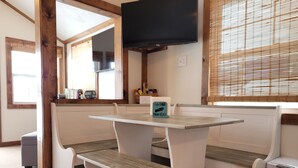 A dining area with a wooden bench and a table, a television mounted on the wall, and a window with blinds. - Sunset Lover's Dream! Lakefront Cottage on Otisco! (Marietta)
