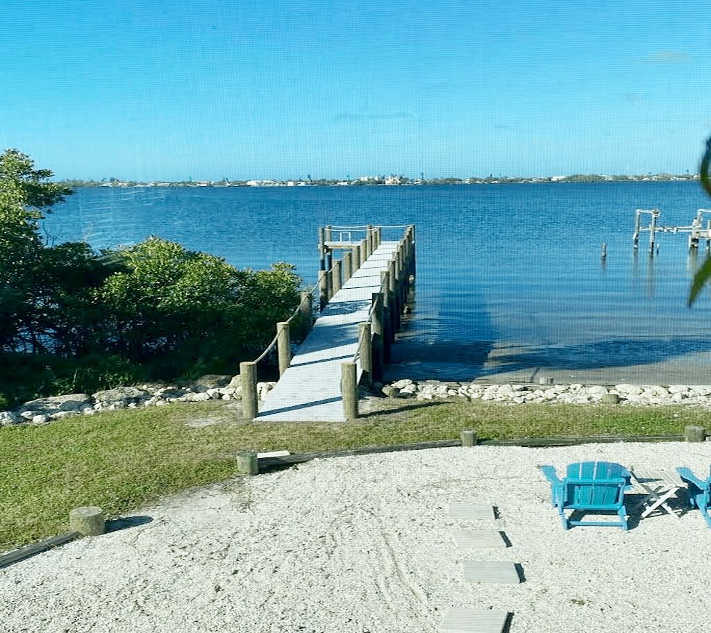Une plage avec une jetée en bois qui s’avance dans des eaux calmes, entourée de verdure et agrémentée de quelques chaises de plage.