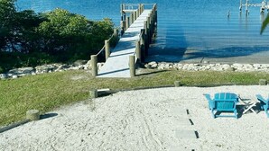Una playa con un muelle de madera que se adentra en aguas tranquilas, rodeada de vegetación y algunas sillas de playa.