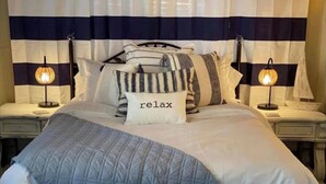 A neatly made bed with a quilted blue blanket, striped pillows, and a pillow with the word "relax" on it. There are two bedside lamps on either side of the bed. - The Comfy Cottage at Possum Kingdom Lake 🚤🐟🌞 (Graford)