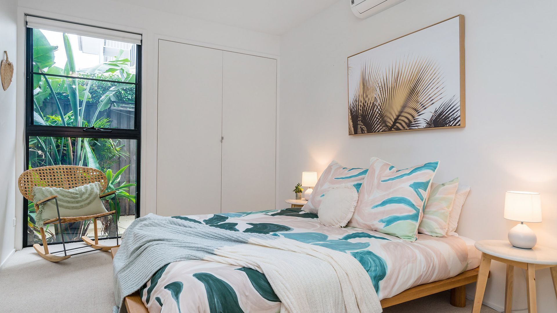 Een slaapkamer met een bed, een stoel, een nachtkastje, een lamp en een raam met uitzicht op groen.