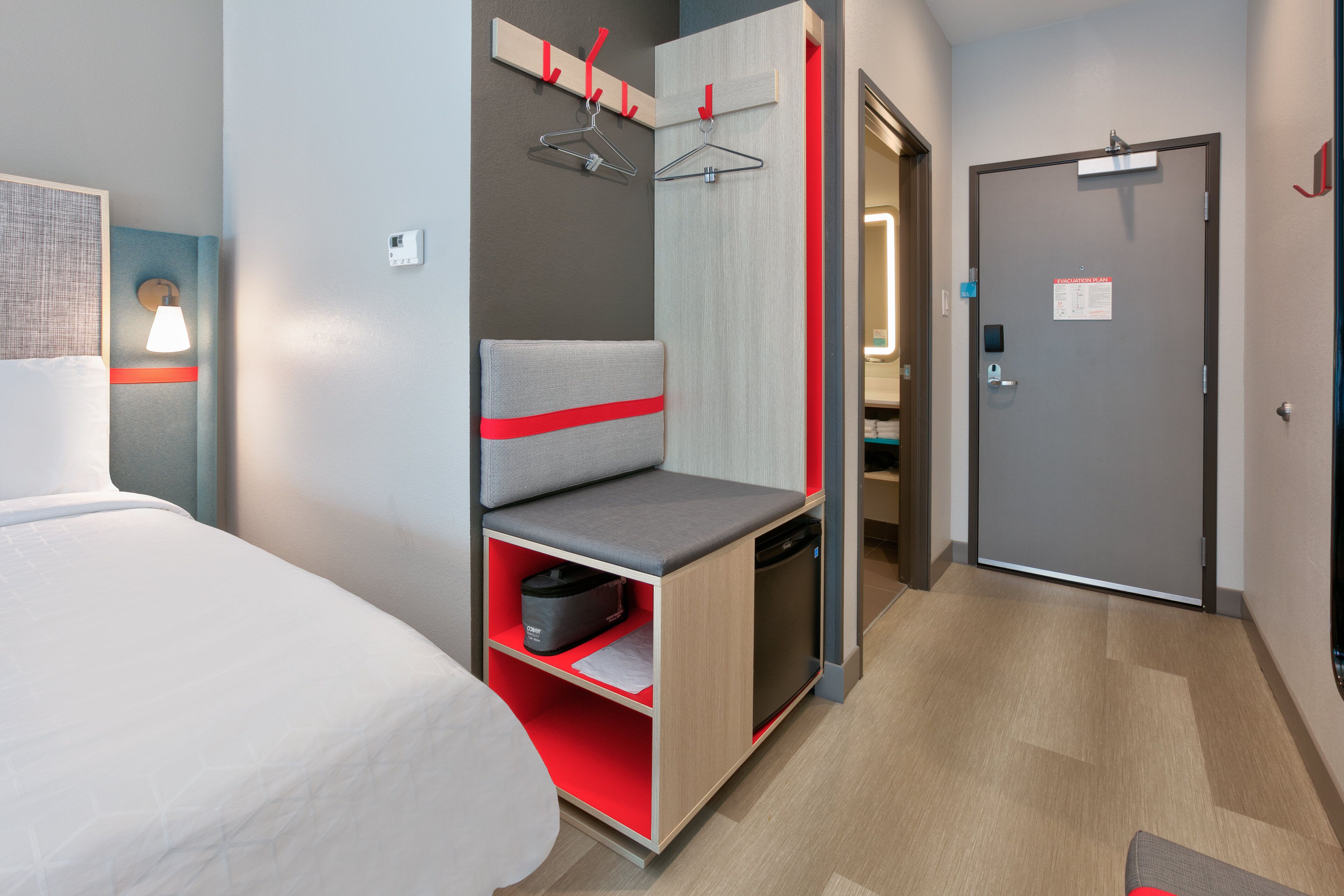 A hotel room with a bed, a seating area with a red cushion, a wardrobe, and a door with a sign.