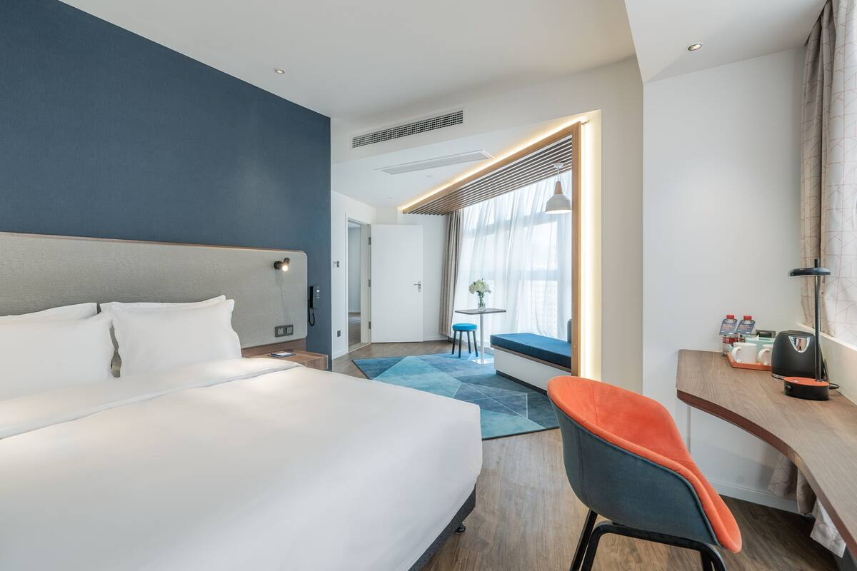 A hotel room with a large bed, a desk with a chair, a small table with a plant, and a window with curtains.