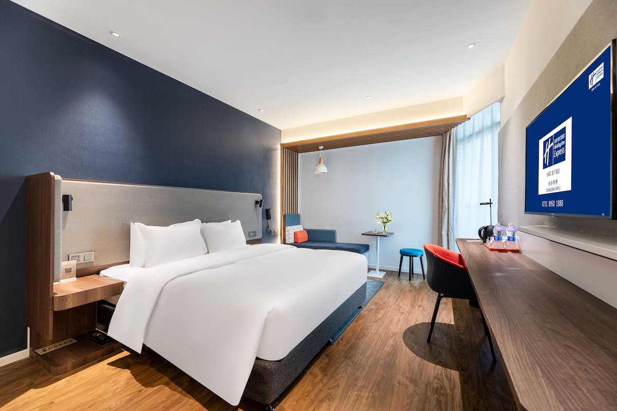 A modern hotel room with a large bed, a desk, a chair, and a TV on the wall.