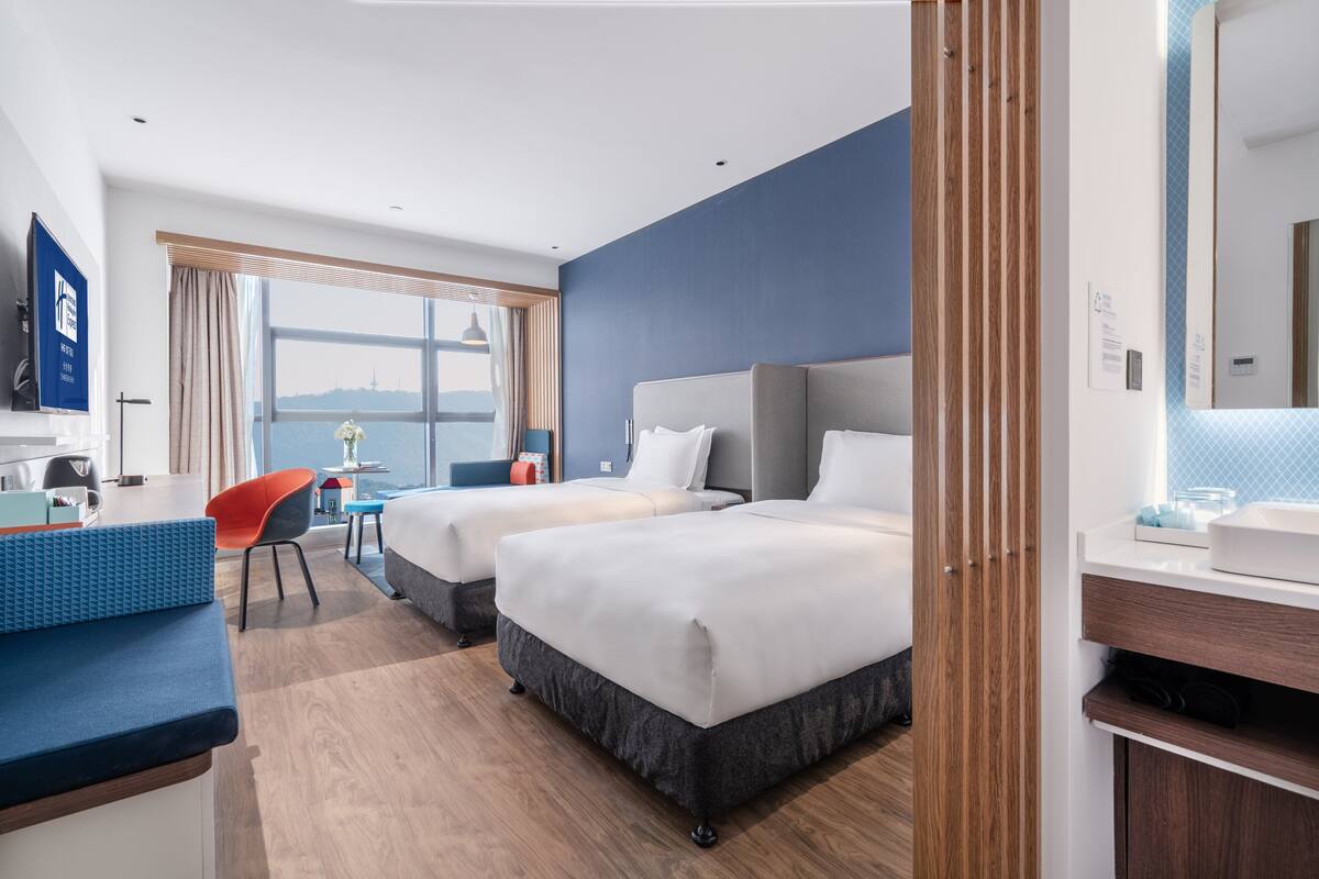 A hotel room with two beds, a desk, a chair, a TV, and a view of the city.
