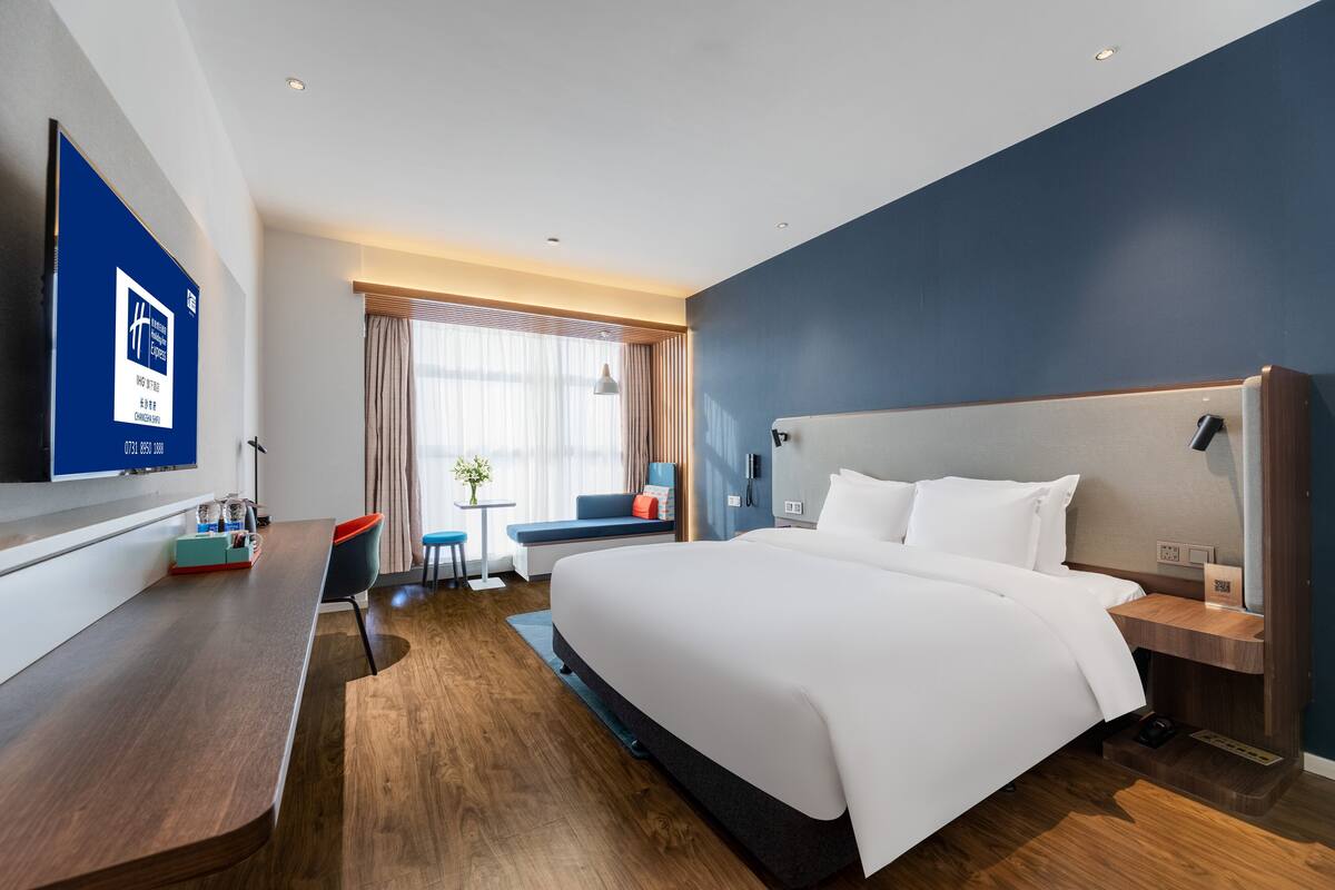 A modern hotel room with a large bed, a desk, a TV, and a window with curtains.