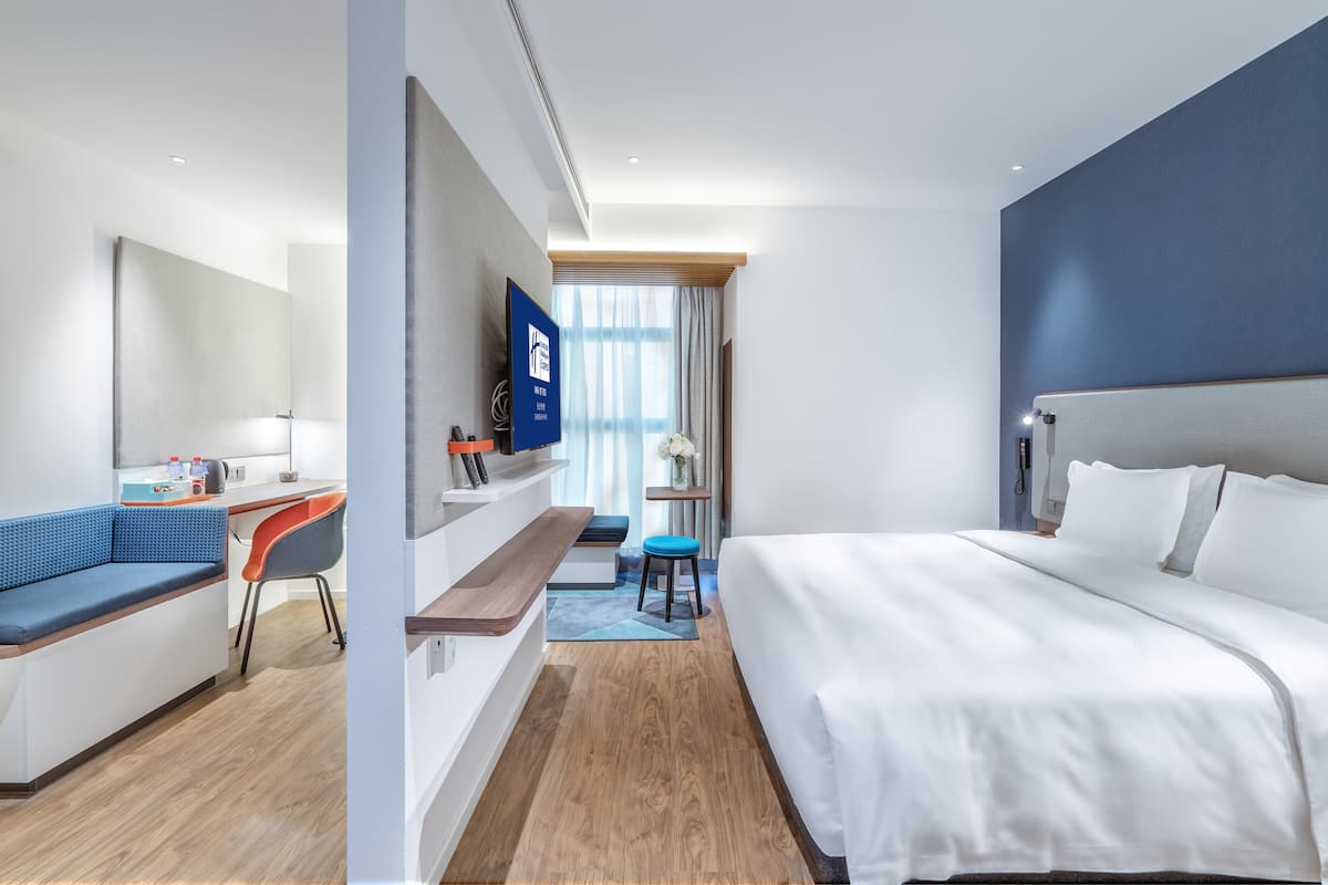 A modern hotel room with a large bed, a desk, a blue sofa, and a TV on the wall.