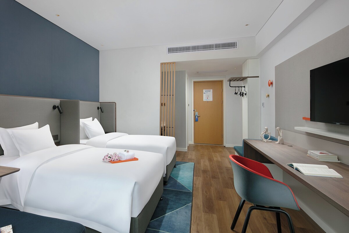 A hotel room with two beds, a desk, a TV, and a bathroom visible through an open door.