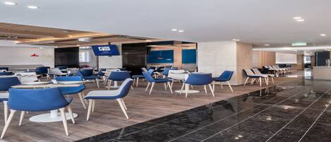 A modern hotel lobby with a mix of seating areas, including tables and chairs, and a reception desk.