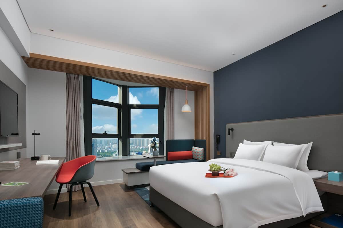 A modern hotel room with a large bed, a desk, a chair, and a TV mounted on the wall.