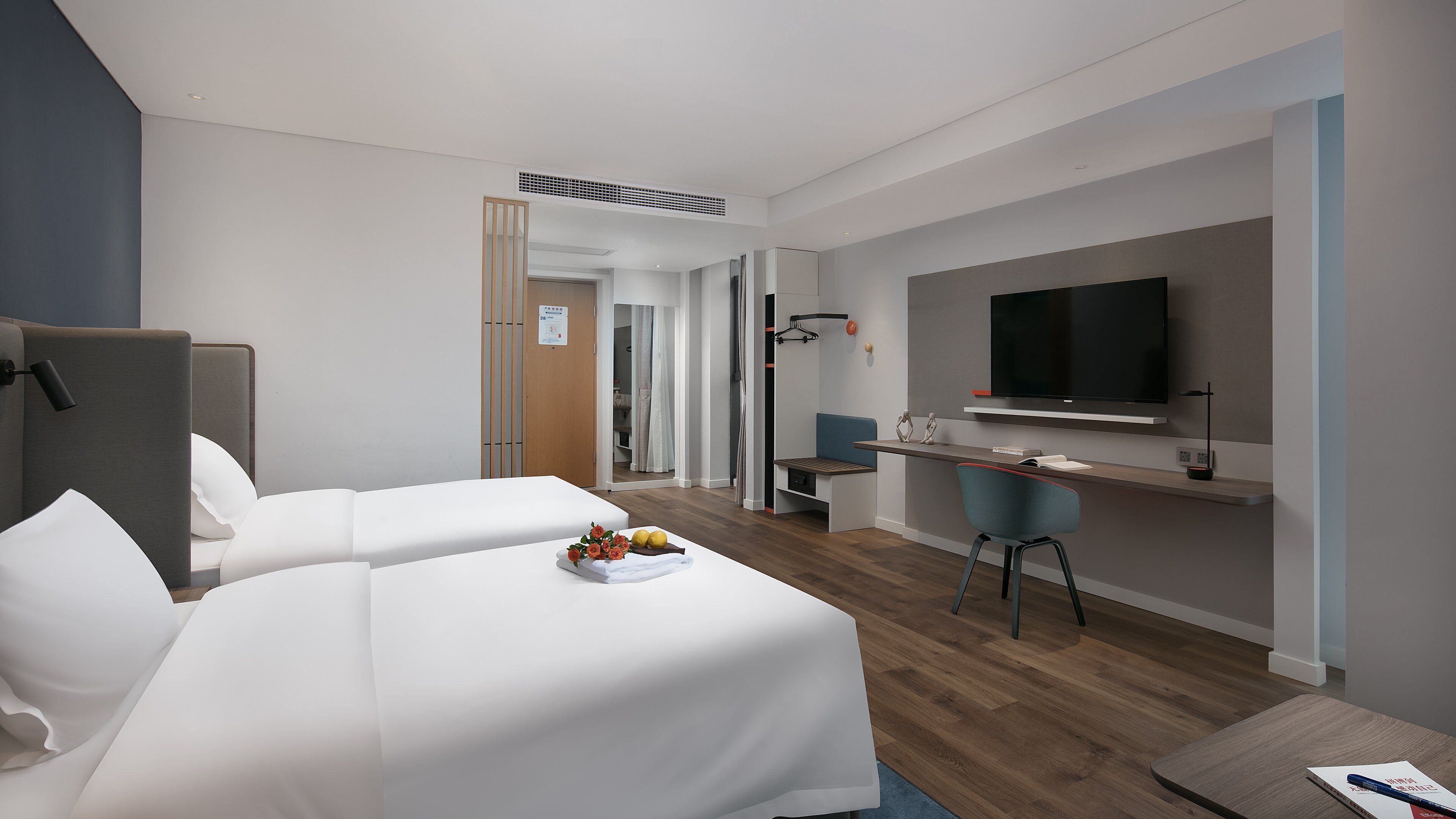 A modern hotel room with two beds, a desk, a TV, and a small seating area.