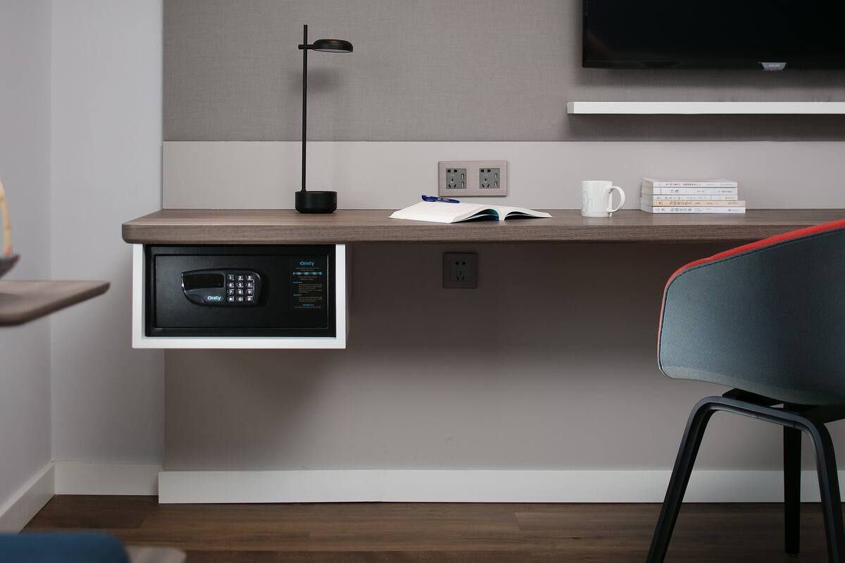 A modern hotel room with a built-in desk, a flat-screen TV mounted on the wall, a desk lamp, and a chair with a red cushion.