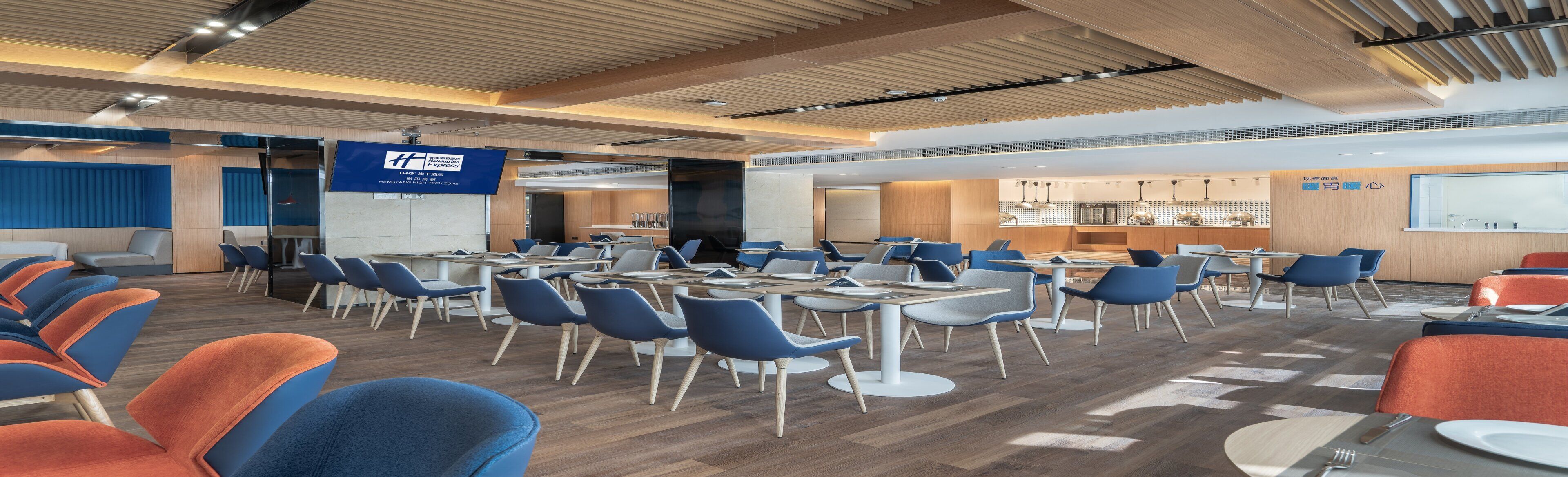 A modern conference room with tables and chairs, a large screen, and a kitchen area in the background.