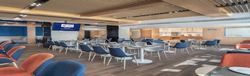 Sala de conferência moderna com mesas, cadeiras, uma tela grande e uma área de cozinha ao fundo.