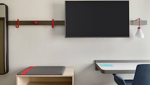A modern room with a flat-screen TV mounted on the wall, a desk with a chair, and a small shelf with a red cushion.