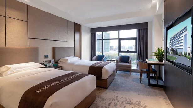 Habitación de hotel con dos camas, un escritorio, una silla, un televisor y un ventanal con vista a la ciudad.
