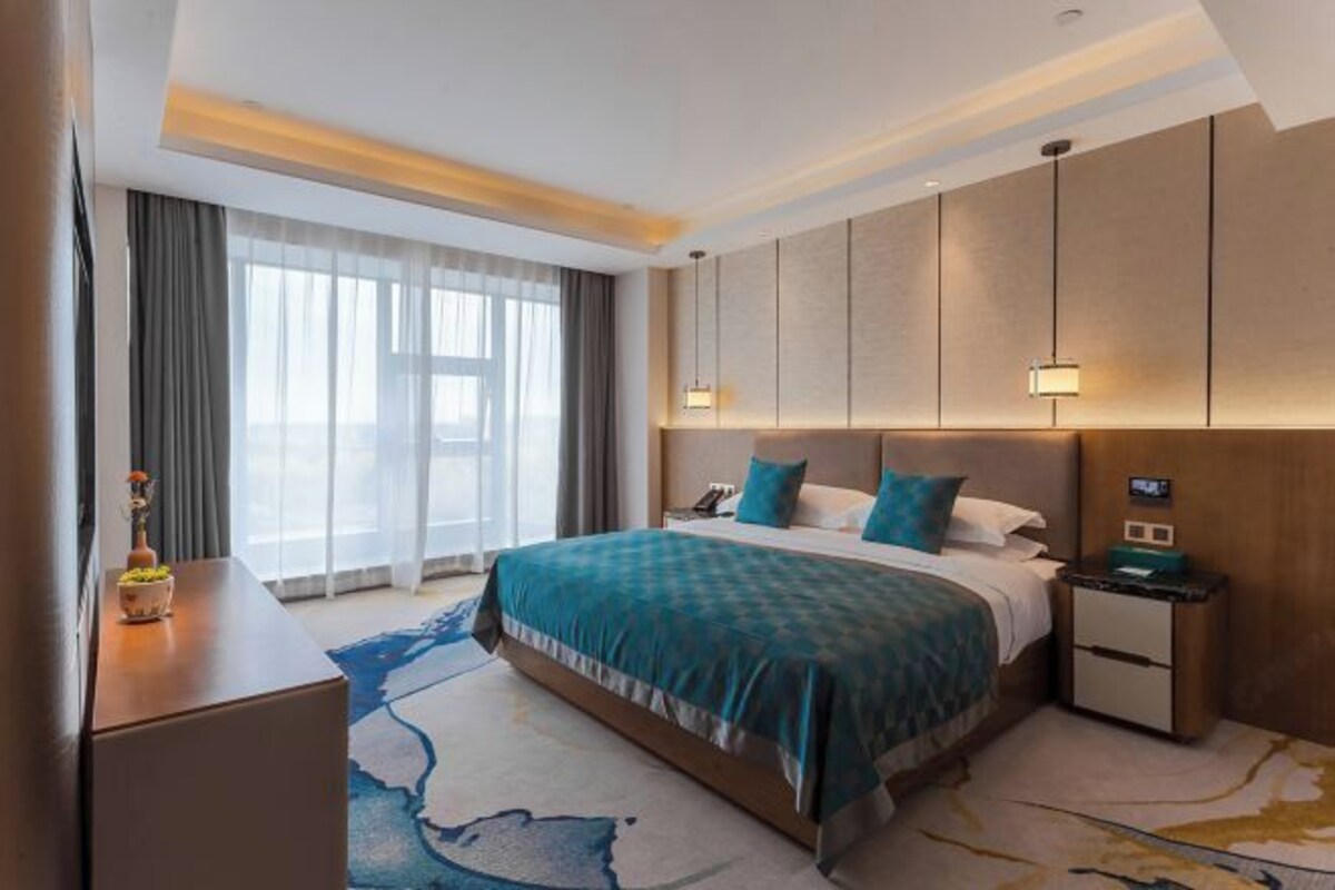 Habitación de hotel moderna con una cama grande, una mesita de noche, un escritorio y vistas a la ciudad a través de amplias ventanas.