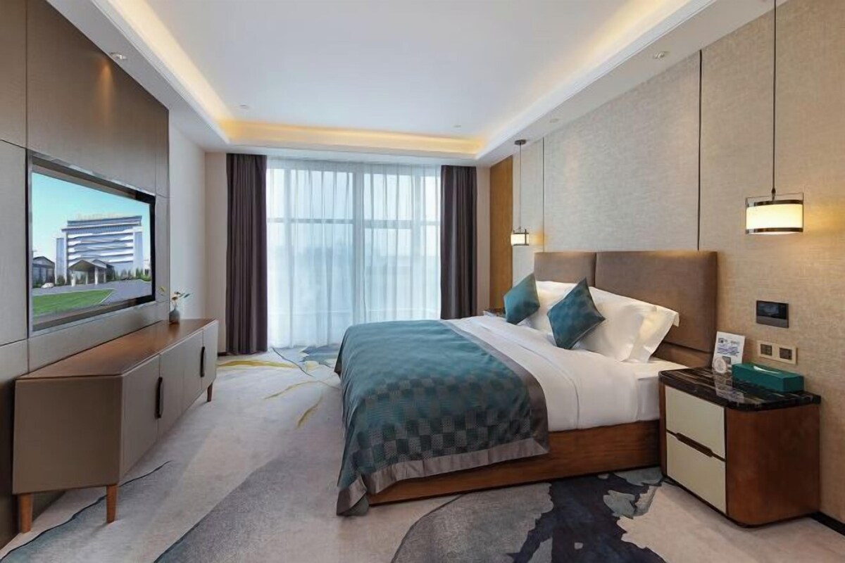 Una habitación de hotel moderna con una cama grande, un televisor de pantalla plana, un escritorio y un amplio ventanal con cortinas.