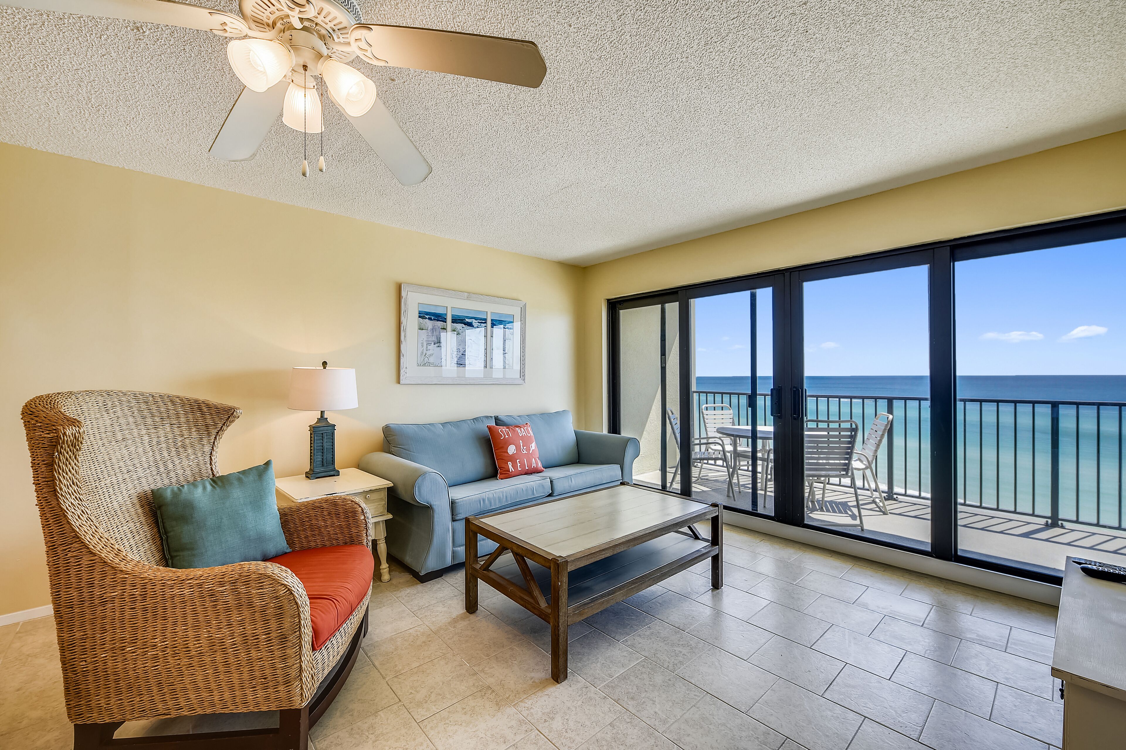 A room with a wicker chair, a sofa, a coffee table, a ceiling fan, a window with a view of the ocean, and a balcony with outdoor furniture.