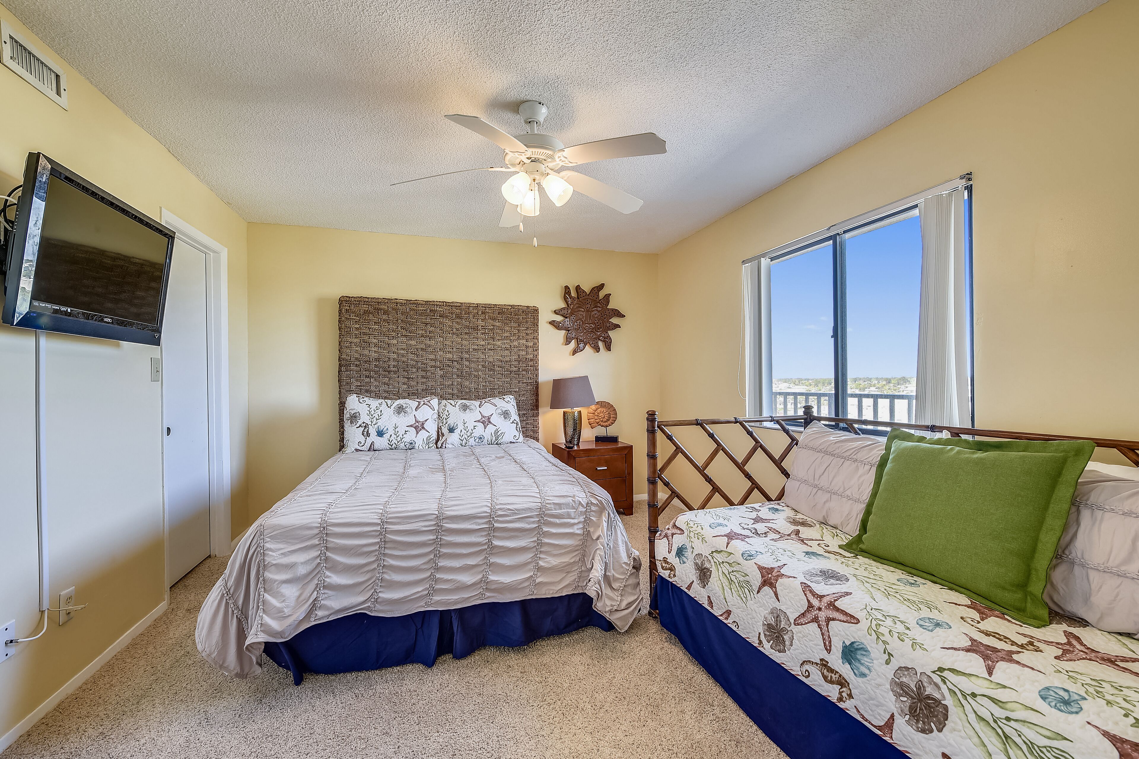A bedroom with a bed, a nightstand, a ceiling fan, a window, and a wall-mounted TV.