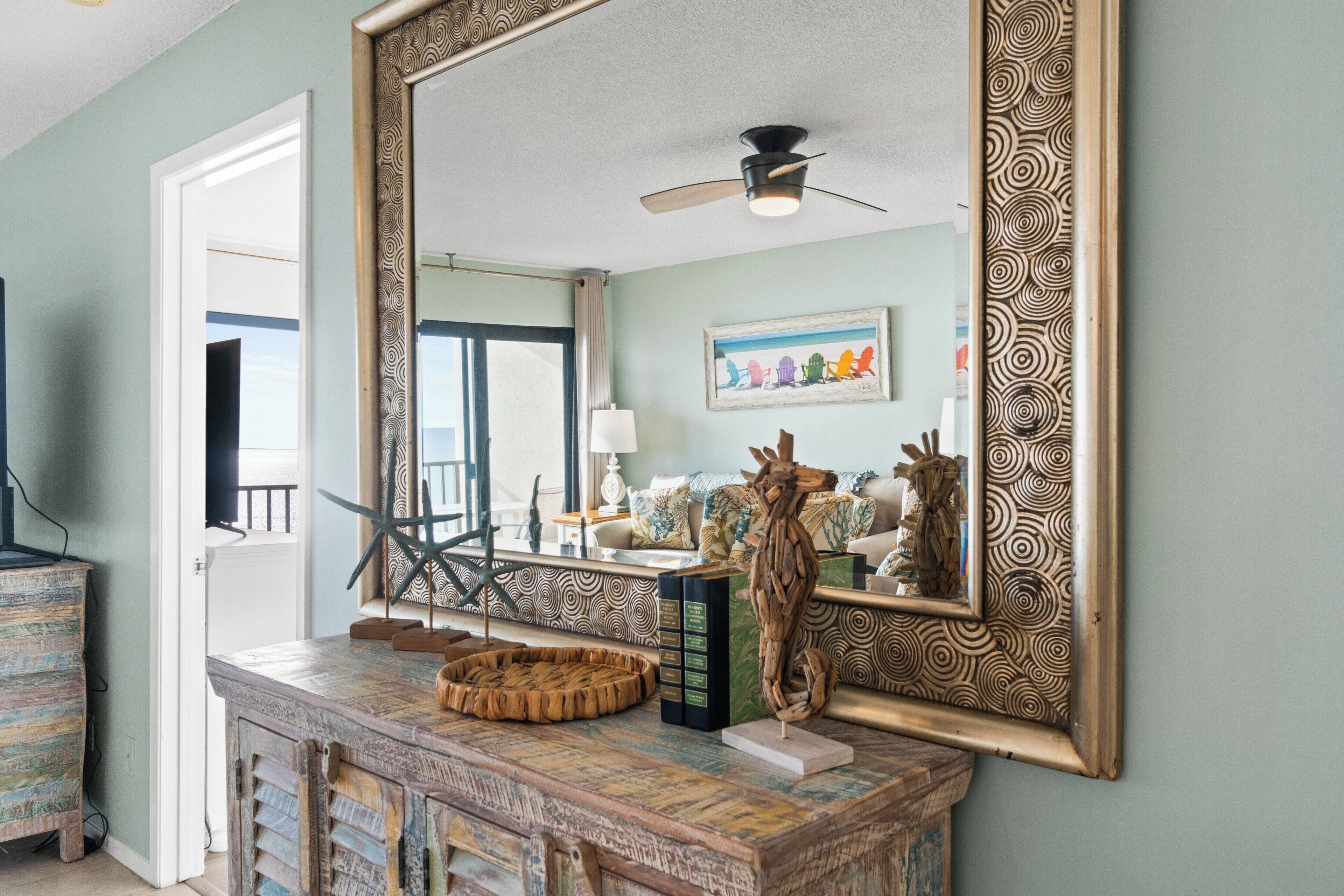 A room with a large mirror, a ceiling fan, a decorative wooden cabinet, and a view of the outdoors through a door.