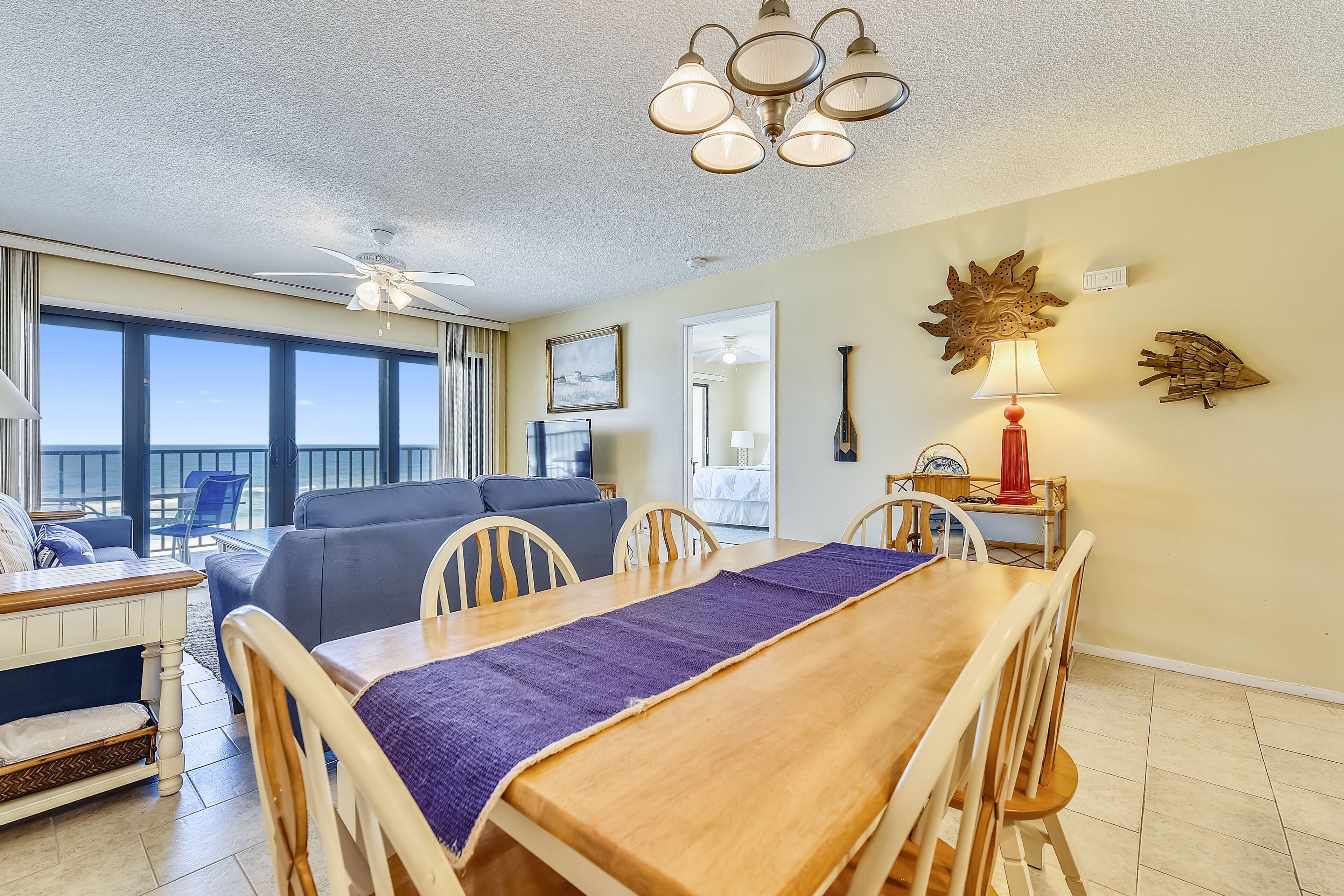 A dining area with a wooden table, chairs, and a purple runner. There is a ceiling fan, a wall-mounted lamp, and a decorative wall piece.