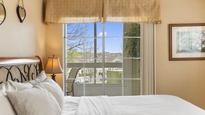 Ein Schlafzimmer mit Bett, Stuhl, Lampe, Fenster mit Blick nach draußen und einem gerahmten Bild an der Wand.