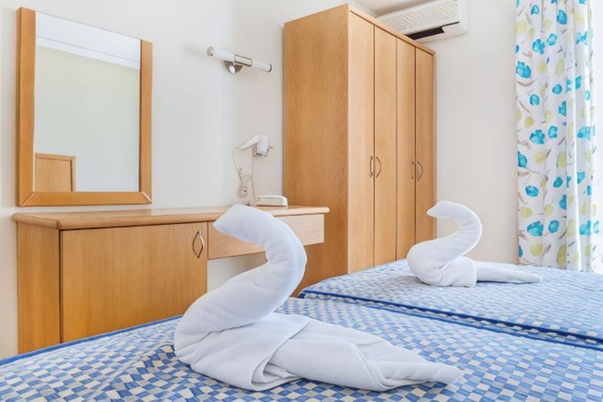 A hotel room with two beds, a wooden wardrobe, a mirror, a towel rack, and a shower curtain with a floral pattern.