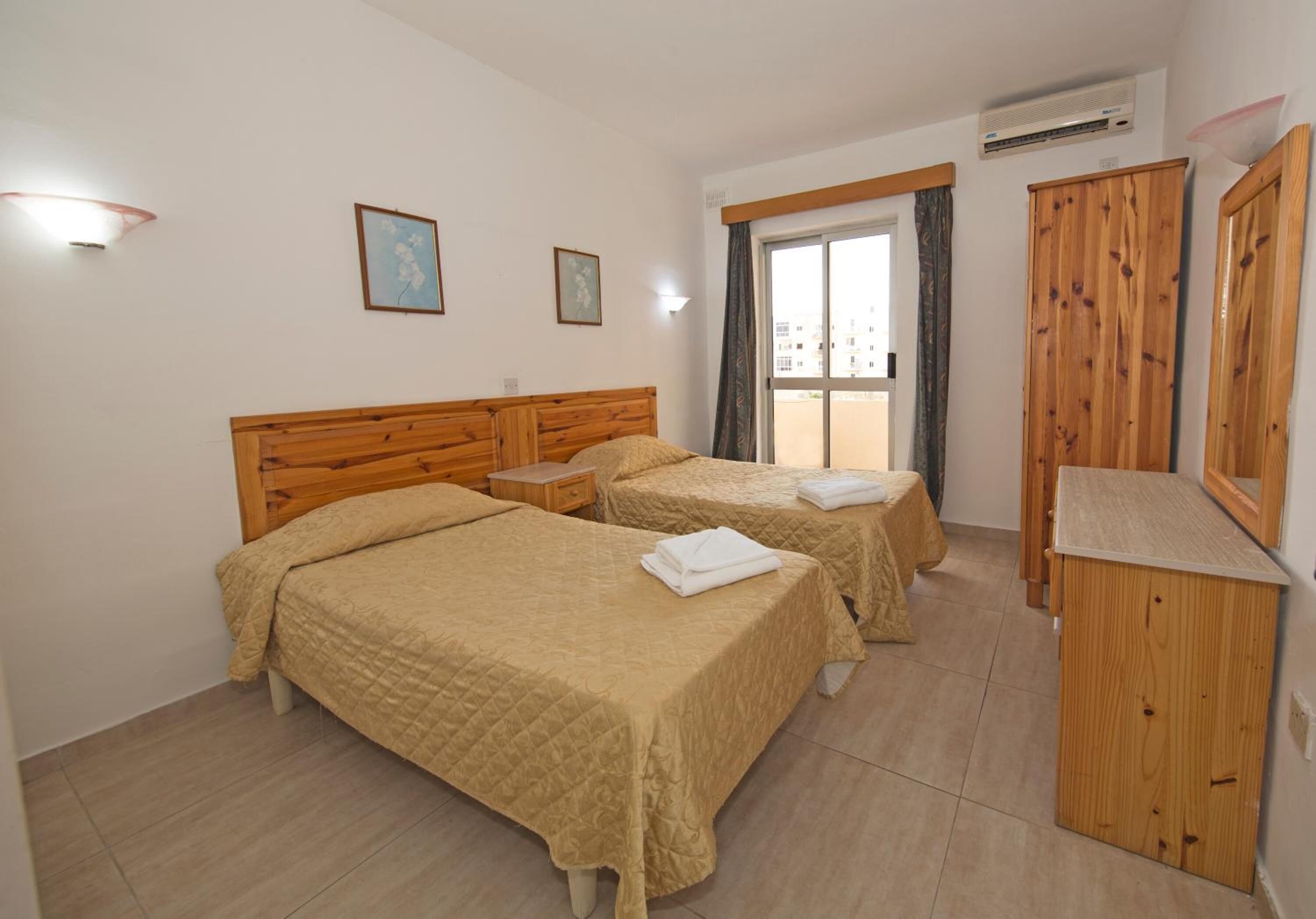 A hotel room with two beds, wooden headboards, a small bedside table, a wooden wardrobe, and a window with curtains.