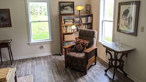 Un cuarto acogedor con piso de madera, una estantería, una silla con cojín, una mesita y dos ventanas que dan al exterior.