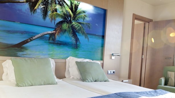 A hotel room with a bed, two pillows, a nightstand, a painting of a palm tree and ocean, and a door. - Nelva (Murcia)