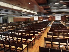 Un salón de conferencias con filas de sillas y mesas, piso de madera e iluminación empotrada en el techo.