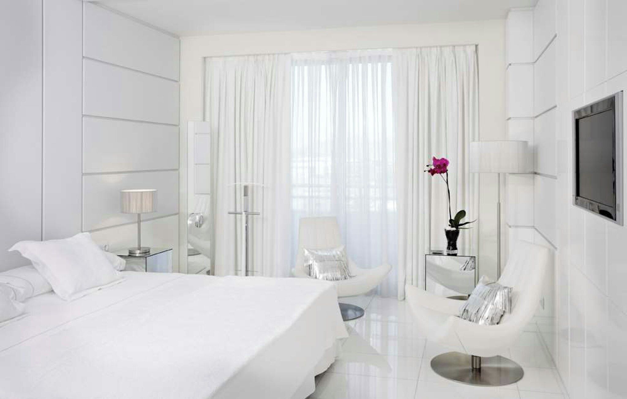 Habitación de hotel moderna con una cama grande, una zona de estar con una silla y una mesita, un televisor montado en la pared y un ventanal con cortinas de encaje.