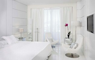 Habitación de hotel moderna con una cama grande, una zona de estar con una silla y una mesita, un televisor montado en la pared y un ventanal con cortinas de encaje.
