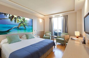 Habitación de hotel con una cama grande, dos sillones, una mesita, un televisor y un cuadro de una playa.