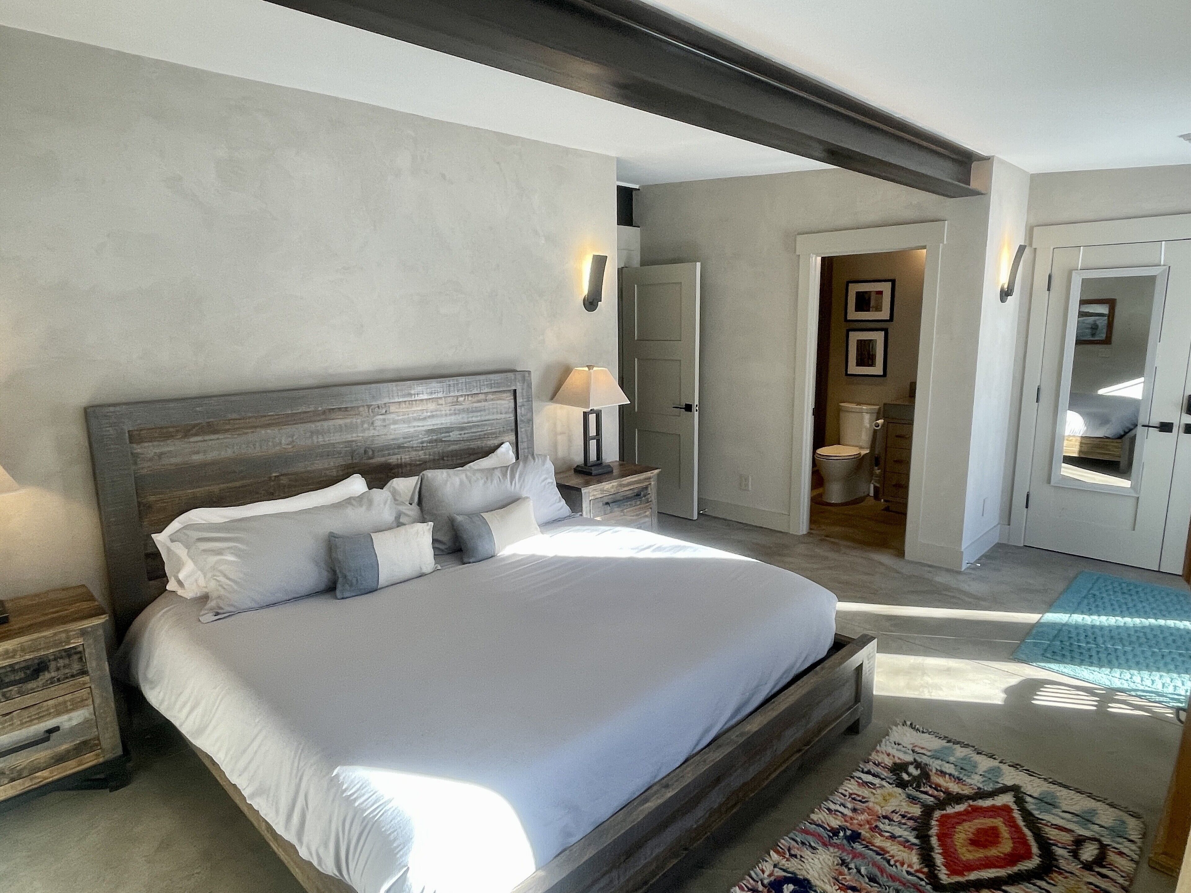 A bedroom with a large bed, a nightstand, a lamp, a door leading to another room, and a patterned rug.