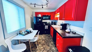 A compact kitchen with red cabinets, a black refrigerator, a dining table with chairs, and a window with blinds.