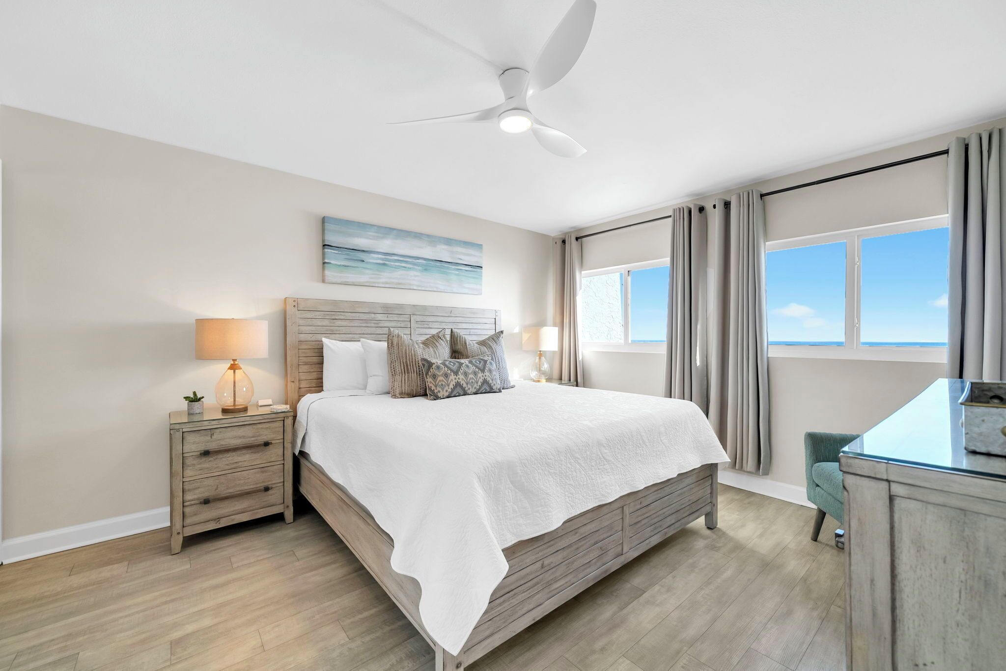 A bedroom with a bed, nightstands, a desk, a ceiling fan, a window with curtains, and a painting on the wall.