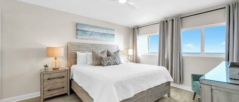 A bedroom with a bed, nightstands, a desk, a ceiling fan, a window with curtains, and a painting on the wall.