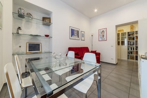 A modern living room with a glass dining table, white chairs, a red sofa, and a bookshelf.