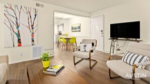 A modern living room with a wooden floor, a dining area with yellow chairs, a TV mounted on the wall, and a painting on the wall. - Walk to Golf Course! Near Lake + Pool w Parking (Austin)